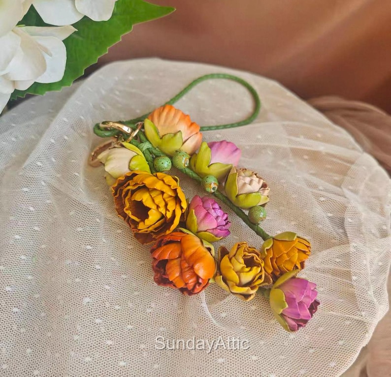 May include: A floral keychain featuring a cluster of colorful, handcrafted flowers in shades of yellow, orange, and pink. The flowers are attached to a green leather cord with a gold-colored clasp. The text "SundayAttic" is visible.