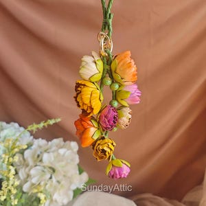 May include: A colorful floral ornament featuring a cluster of fabric flowers in shades of yellow, orange, pink, and white. The flowers are attached to a green braided cord with a gold-colored ring. The text "SundayAttic" is visible at the bottom.