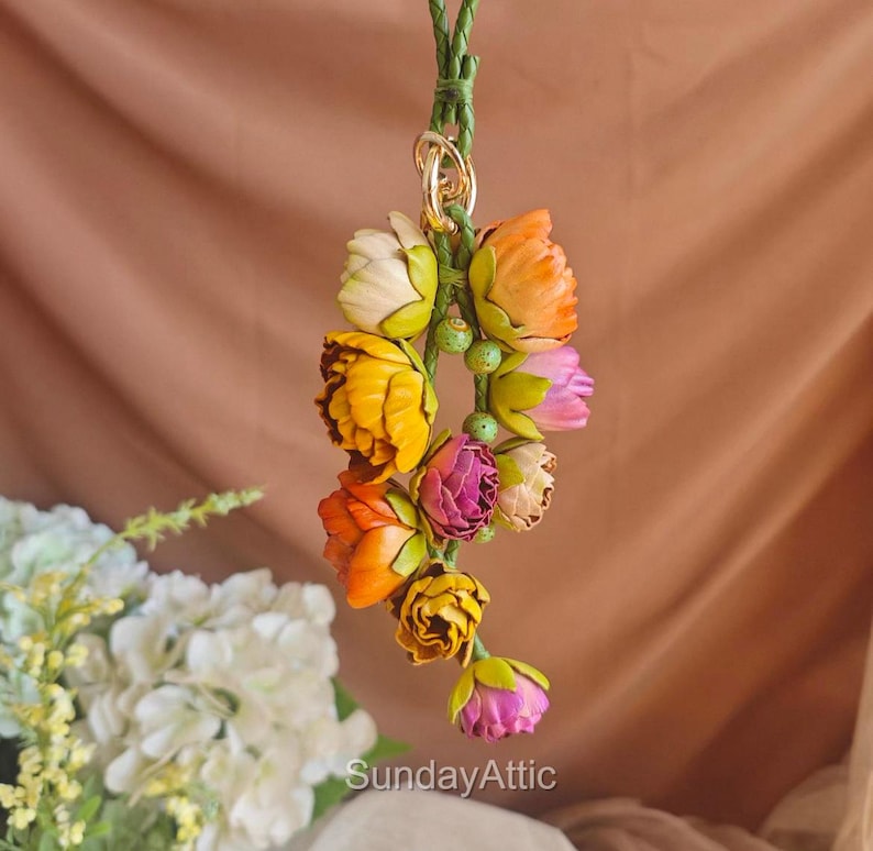 May include: A colorful floral ornament featuring a cluster of fabric flowers in shades of yellow, orange, pink, and white. The flowers are attached to a green braided cord with a gold-colored ring. The text "SundayAttic" is visible at the bottom.