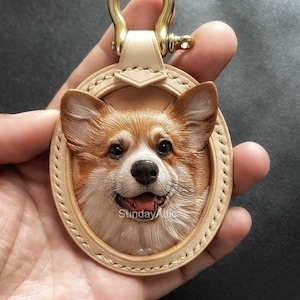 Op de afbeelding: Ovale leren sleutelhanger met een gedetailleerde corgi-gezicht in bruin, wit en beige. Het heeft een goudkleurige metalen sluiting en de tekst "Sunday Attic".