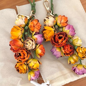 May include: Two floral keychains with a variety of colorful flowers in shades of orange, yellow, and purple. The flowers are attached to green stems and have gold-colored clasps. The keychains are displayed on a cream-colored surface.