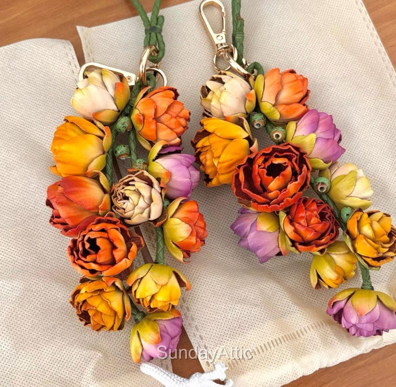 May include: Two floral keychains with a variety of colorful flowers in shades of orange, yellow, and purple. The flowers are attached to green stems and have gold-colored clasps. The keychains are displayed on a cream-colored surface.