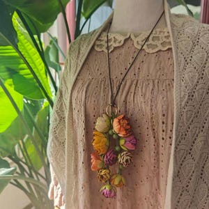May include: A floral pendant necklace with a cluster of colorful fabric flowers in shades of orange, yellow, pink, and purple. The necklace hangs from a dark brown cord and a gold-colored ring. The text "SundayAttic" is visible.