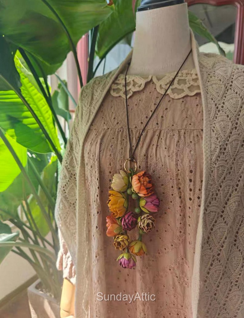 May include: A floral pendant necklace with a cluster of colorful fabric flowers in shades of orange, yellow, pink, and purple. The necklace hangs from a dark brown cord and a gold-colored ring. The text "SundayAttic" is visible.