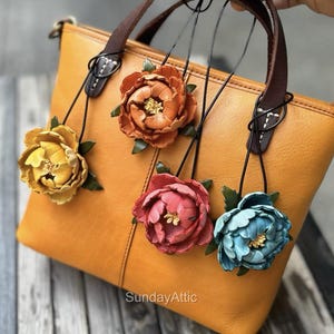 Può includere: Una borsa in pelle giallo senape con manici marroni e cuciture decorative. Sono attaccati cinque ciondoli a forma di fiore in pelle gialla, arancione, rosa e blu. I ciondoli sono sospesi da cordini neri. Il testo "SundayAttic" è visibile.