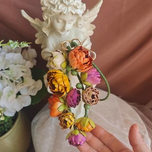 May include: A colorful floral bouquet featuring orange, yellow, and purple flowers. The bouquet is attached to a gold-colored ring. The text "SundayAttic" is visible at the bottom of the image. A white angel statue is in the background.