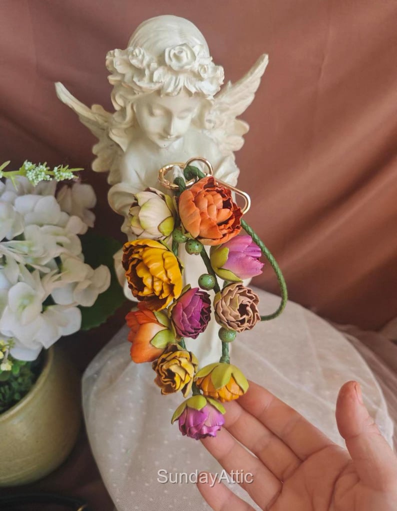 May include: A colorful floral bouquet featuring orange, yellow, and purple flowers. The bouquet is attached to a gold-colored ring. The text "SundayAttic" is visible at the bottom of the image. A white angel statue is in the background.