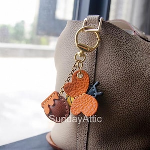May include: A gold-coloured keychain with a honey pot, heart, and bee charm. The honey pot is brown with a tan top, the heart is orange, and the bee has yellow and black stripes. The text "SundayAttic" is visible.