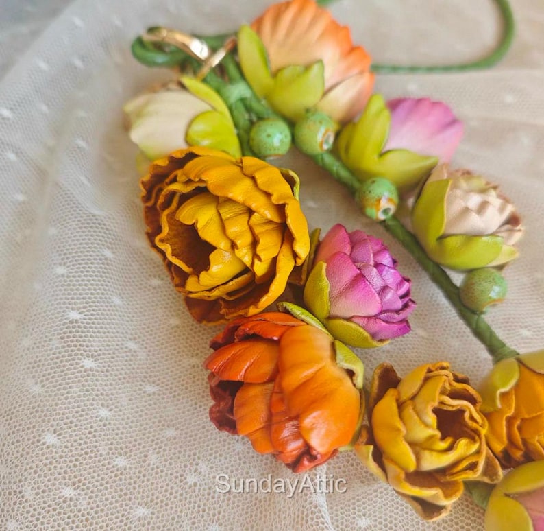 May include: A floral accessory featuring handcrafted leather flowers in shades of yellow, orange, pink, and cream. The flowers are attached to a green stem, creating a vibrant and artistic design. The text "Sunday Attic" is visible.