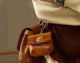 May include: A small, tan leather pouch with a brass button closure and a gold chain. The pouch is attached to a larger brown leather bag. The text "Sunday Attic" is printed on the pouch.