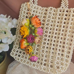 May include: A cream-colored beaded handbag with a floral charm. The charm features colorful fabric flowers in shades of orange, yellow, pink, and purple. The text "Sunday Attic" is visible at the bottom of the image.