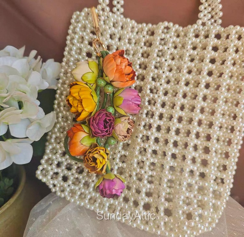 May include: A cream-colored beaded handbag with a floral charm. The charm features colorful fabric flowers in shades of orange, yellow, pink, and purple. The text "Sunday Attic" is visible at the bottom of the image.