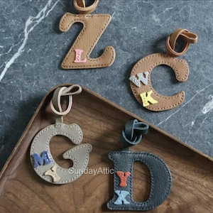 May include: Leather letter keychains in brown, gray, and dark gray. The letters Z, C, G, and D are visible, each with smaller letters stitched on. The keychains are on a wooden tray and a gray marble surface.