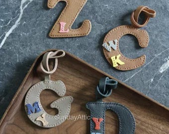 May include: Leather letter keychains in brown, gray, and dark gray. The letters Z, C, G, and D are visible, each with smaller letters stitched on. The keychains are on a wooden tray and a gray marble surface.