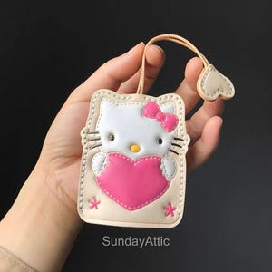 May include: A beige and white Hello Kitty-themed keychain wallet with a pink heart and bow. The wallet has a stitched border and a small loop with a heart-shaped charm. The text "SundayAttic" is visible.