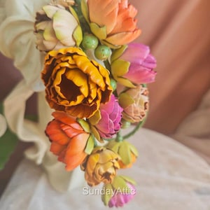 May include: A close-up of a floral arrangement featuring vibrant, colorful flowers. The arrangement includes orange, yellow, and pink blooms with green accents. The flowers are clustered together, creating a visually appealing display. The text "SundayAttic" is visible.