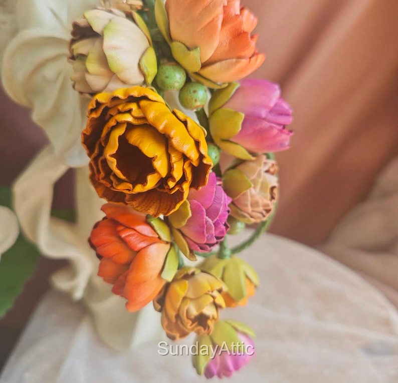 May include: A close-up of a floral arrangement featuring vibrant, colorful flowers. The arrangement includes orange, yellow, and pink blooms with green accents. The flowers are clustered together, creating a visually appealing display. The text "SundayAttic" is visible.