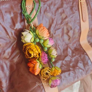 May include: A leather bag charm featuring a cluster of colorful, handcrafted flowers in shades of yellow, orange, pink, and white. The flowers are attached to a leather strap with a green braided cord. The text "Sunday Attic" is visible.