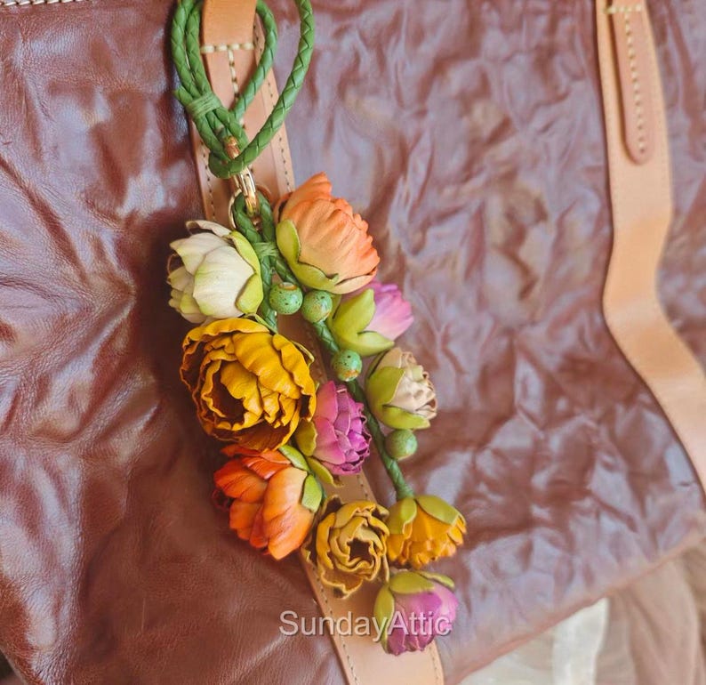 May include: A leather bag charm featuring a cluster of colorful, handcrafted flowers in shades of yellow, orange, pink, and white. The flowers are attached to a leather strap with a green braided cord. The text "Sunday Attic" is visible.
