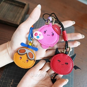 May include: Three colorful leather keychains with cartoon faces. One is orange with goggles, one is pink with a cloud, and the last is red with devil horns and wings. Each keychain has a different colored cord.