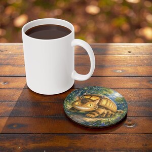 Pu&ograve; includere: Una tazza da caff&egrave; in ceramica bianca piena di liquido scuro &egrave; appoggiata su un tavolo di legno accanto a un sottobicchiere rotondo. Il sottobicchiere presenta un gatto dei cartoni animati con un corpo a strisce e un volto sorridente.
