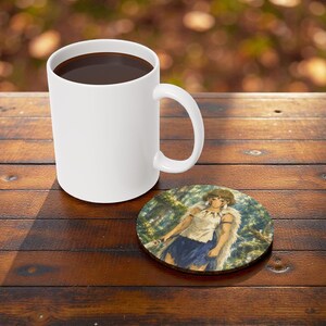May include: A white ceramic coffee mug filled with dark liquid sits on a wooden table next to a round coaster. The coaster features a colorful illustration of a person in a forest setting.