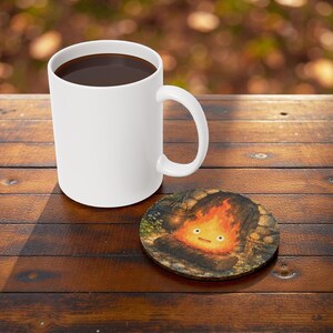 Pu&ograve; includere: Una tazza di ceramica bianca piena di liquido scuro &egrave; appoggiata su un tavolo di legno accanto a un sottobicchiere rotondo. Il sottobicchiere presenta un'illustrazione a cartoni animati di un fal&ograve; con una faccia sorridente.