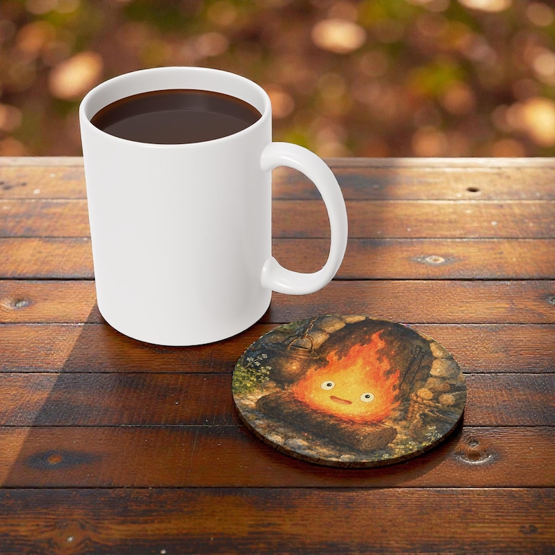 Pu&ograve; includere: Una tazza di ceramica bianca piena di liquido scuro &egrave; appoggiata su un tavolo di legno accanto a un sottobicchiere rotondo. Il sottobicchiere presenta un'illustrazione a cartoni animati di un fal&ograve; con una faccia sorridente.