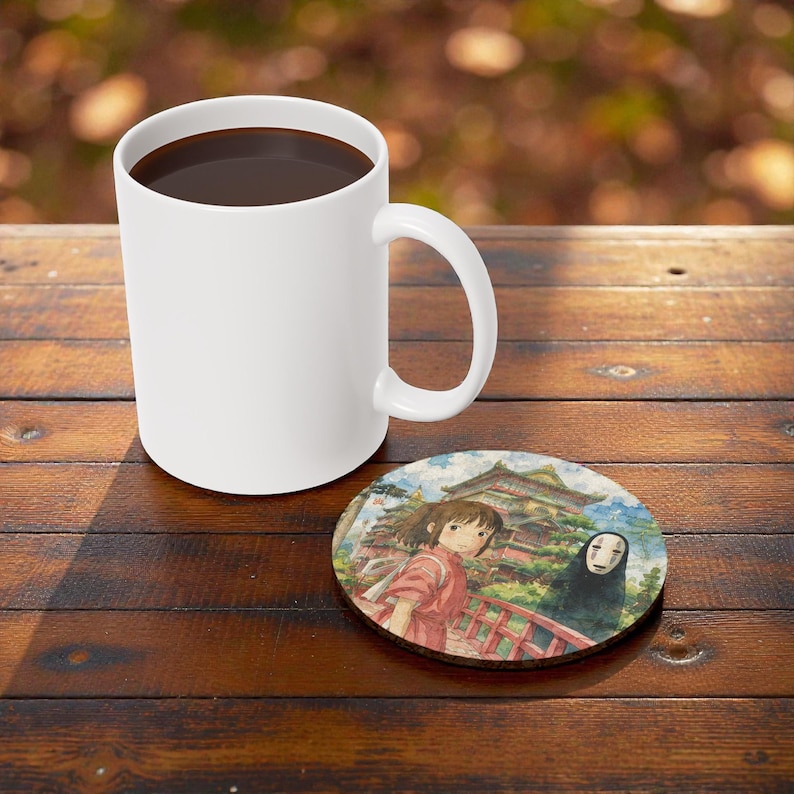 Pu&ograve; includere: Una tazza da caff&egrave; in ceramica bianca piena di liquido scuro, accanto a un sottobicchiere rotondo con una scena anime con una ragazza e una figura mascherata. Il sottobicchiere ha un aspetto di legno ed &egrave; posto su una superficie di legno.