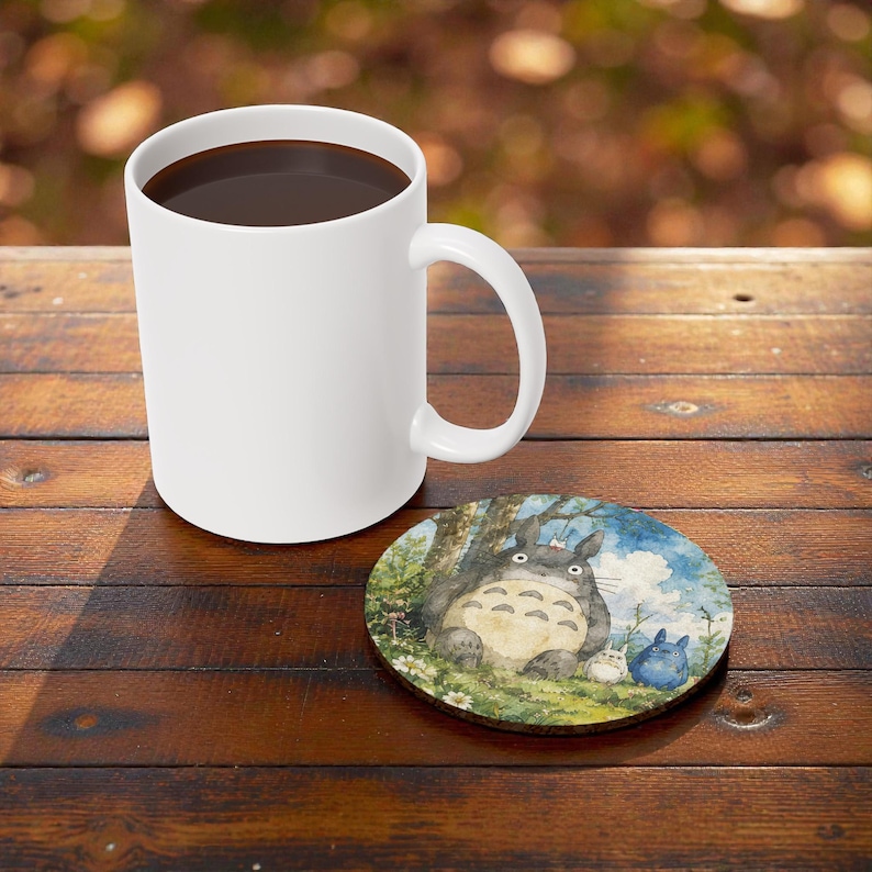 Pu&ograve; includere: Una tazza da caff&egrave; in ceramica bianca piena di liquido scuro &egrave; appoggiata su un tavolo di legno accanto a un sottobicchiere rotondo. Il sottobicchiere presenta un'illustrazione di un grande personaggio grigio e peloso con creature blu e bianche pi&ugrave; piccole.