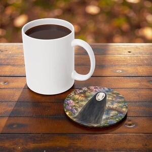 May include: A white ceramic coffee mug filled with dark liquid sits on a wooden table next to a round coaster. The coaster features a watercolor illustration of a figure in a black cloak with a white mask, set against a floral and bridge background.