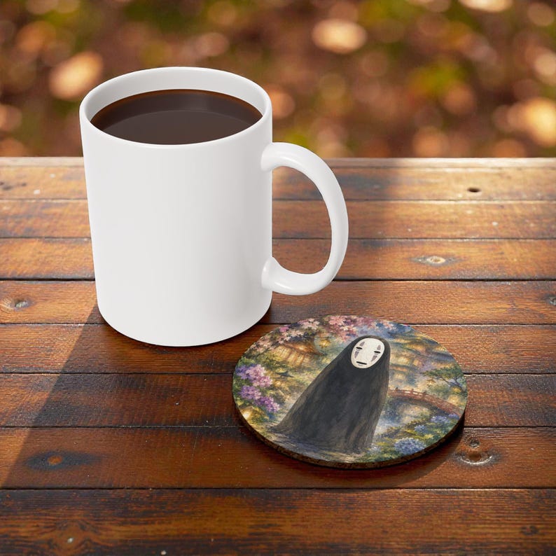 May include: A white ceramic coffee mug filled with dark liquid sits on a wooden table next to a round coaster. The coaster features a watercolor illustration of a figure in a black cloak with a white mask, set against a floral and bridge background.