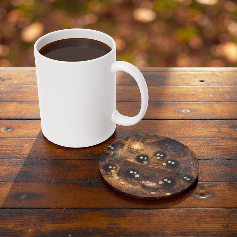 Pu&ograve; includere: Una tazza da caff&egrave; in ceramica bianca piena di liquido scuro &egrave; appoggiata su un tavolo di legno accanto a un sottobicchiere rotondo. Il sottobicchiere presenta un'illustrazione colorata di piccole creature scure e rotonde con grandi occhi.