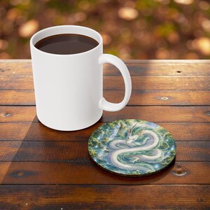 May include: A white ceramic mug filled with dark liquid sits on a wooden table next to a round coaster. The coaster features a detailed illustration of a white dragon with green accents, set against a backdrop of blue and green foliage.