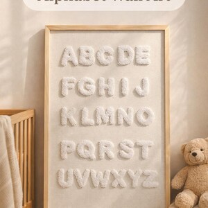 May include: Soft, textured alphabet wall art in a wooden frame. The letters A through Z are displayed in a soft, plush material against a neutral background. A teddy bear and crib are visible in the background.