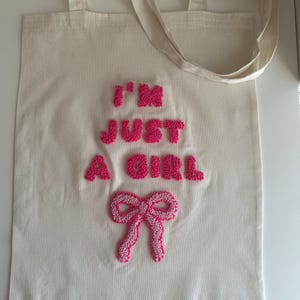 Op de afbeelding: Crèmekleurige draagtas met de tekst "I'M JUST A GIRL" in roze, getextureerde letters. Een roze strik is onder de tekst geborduurd. De tas heeft twee crèmekleurige handvatten.