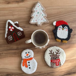 May include: A collection of handmade Christmas-themed coasters. Designs include a gingerbread house, Christmas tree, snowman with an orange scarf, a penguin wearing a Santa hat, and a reindeer with a red scarf. A cup of coffee sits in the center.