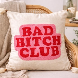 Può includere: Cuscino decorativo color crema con la scritta "BAD BITCH CLUB" in lettere rosse su sfondo rosa. Il cuscino ha nappe su ogni angolo ed è posto su una sedia di vimini.
