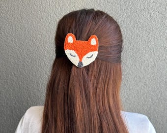 May include: A decorative hair clip featuring a fox design. The clip is orange, white, and black, with a textured appearance. The fox has closed eyes, suggesting a sleeping or relaxed state. The clip is attached to brown hair.