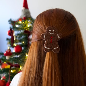 May include: A brown gingerbread man hair clip with white accents and a red button detail is attached to long, auburn hair. A Christmas tree with ornaments is visible in the background, suggesting a holiday theme.