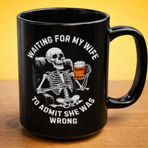 May include: Black ceramic mug with a humorous skeleton graphic. The skeleton is seated, holding a beer, with the text "WAITING FOR MY WIFE TO ADMIT SHE WAS WRONG" in white. The mug has a large handle and is suitable for hot or cold beverages.