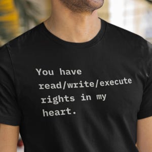 Puede incluir: Camiseta negra con el texto blanco "You have read/write/execute rights in my heart". La camiseta de cuello redondo parece estar hecha de un material suave. El texto está centrado en la parte delantera de la camiseta.