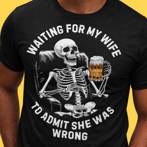 May include: Black t-shirt featuring a skeleton in a chair holding a beer mug. The text on the shirt reads "WAITING FOR MY WIFE TO ADMIT SHE WAS WRONG" in white, curved around the skeleton.