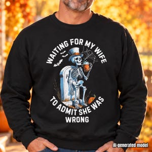 May include: A black crewneck sweatshirt featuring a graphic of a skeleton wearing a top hat, sitting on a tombstone while holding a glass of beer. The text reads "WAITING FOR MY WIFE TO ADMIT SHE WAS WRONG" with bats, a spiderweb, and a pumpkin.