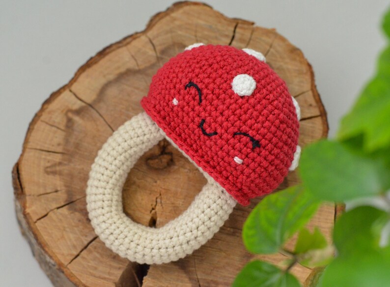May include: A crocheted mushroom toy with a red cap and white spots. The mushroom has a white crocheted ring for a stem and a smiling face.
