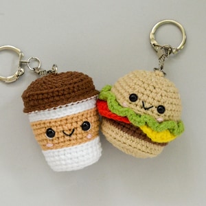 May include: Two crocheted keychains: a coffee cup and a hamburger. The coffee cup is brown and tan with a white base. The hamburger has a tan bun, red tomato, green lettuce, and yellow cheese. Both have smiling faces.