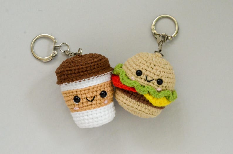 May include: Two crocheted keychains: a coffee cup and a hamburger. The coffee cup is brown and tan with a white base. The hamburger has a tan bun, red tomato, green lettuce, and yellow cheese. Both have smiling faces.