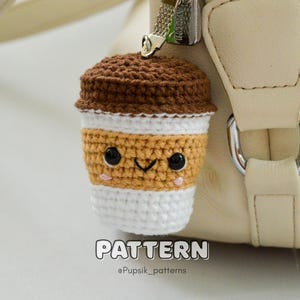 May include: A crocheted coffee cup charm with a smiling face, attached to a beige handbag. The charm features a brown top, a tan middle section, and a white bottom. The word "PATTERN" is displayed at the bottom.