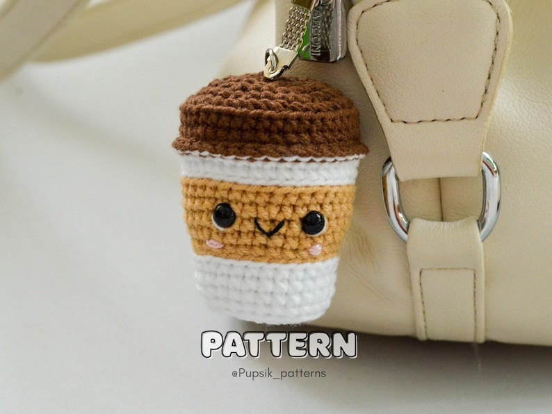 May include: A crocheted coffee cup charm with a smiling face, attached to a beige handbag. The charm features a brown top, a tan middle section, and a white bottom. The word "PATTERN" is displayed at the bottom.