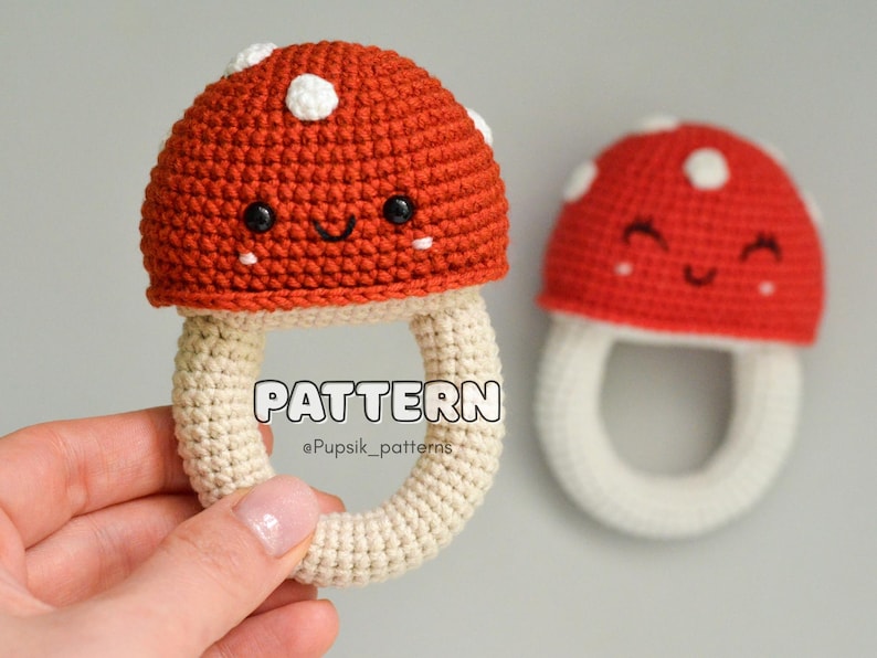 May include: A crocheted mushroom toy with a white stem and a red cap with white dots. The mushroom has a smiling face and is a rattle toy.